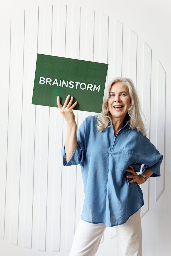 Cheerful senior woman in blue shirt holding a green 'Brainstorm' sign indoors, promoting ideas.